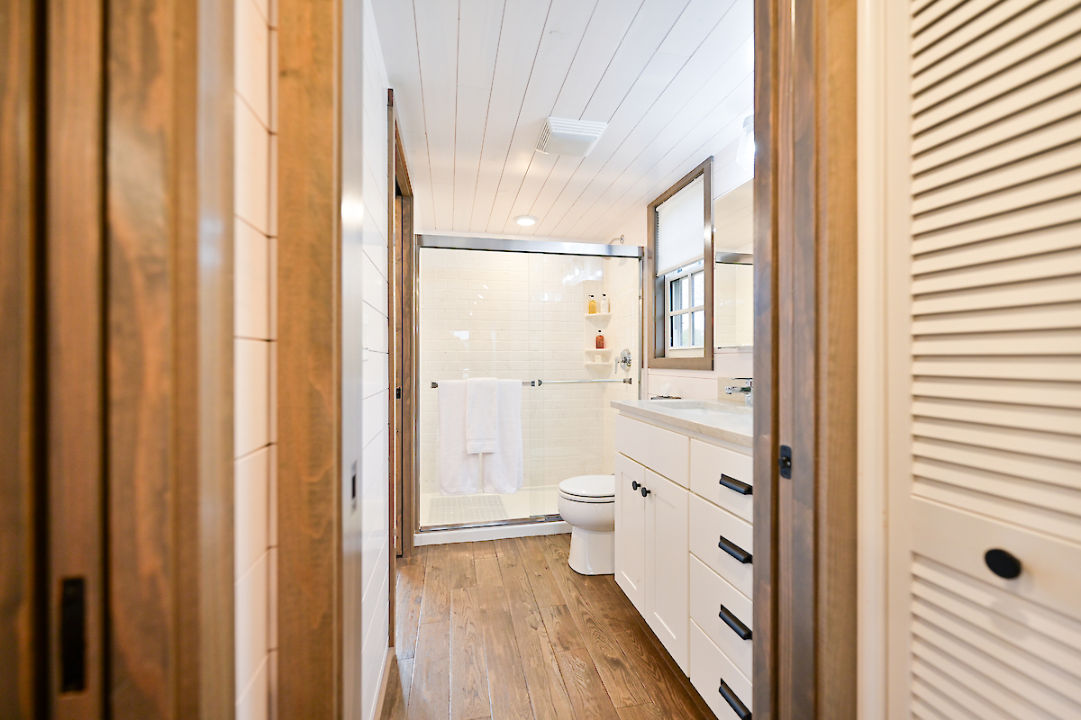 The image shows a modern bathroom with wooden floors, a shower with glass doors, a white vanity with drawers, and a toilet, ending the sentence.
