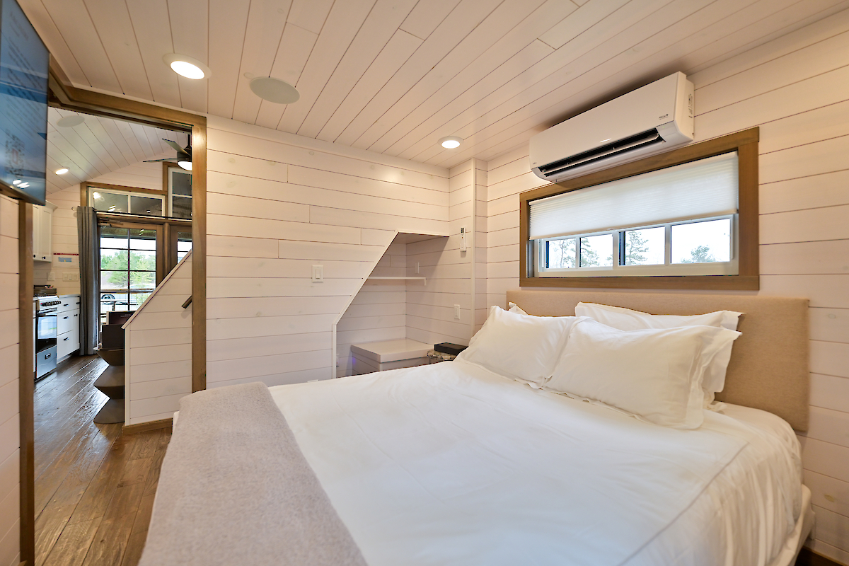 This image shows a cozy bedroom with a double bed, white walls, air conditioning, small windows, and view into a modern kitchen area.