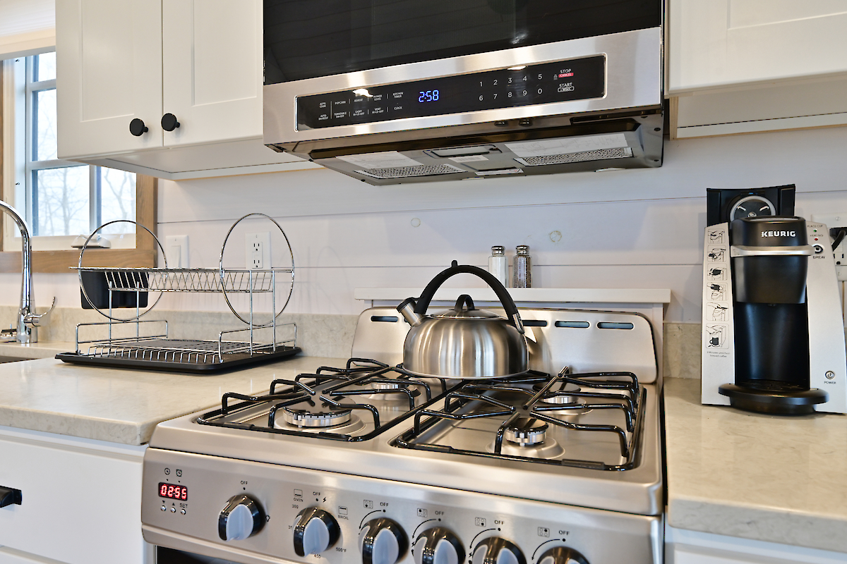 A kitchen with a stovetop, kettle, dish rack, microwave, spice shakers, and a coffee machine on a countertop.