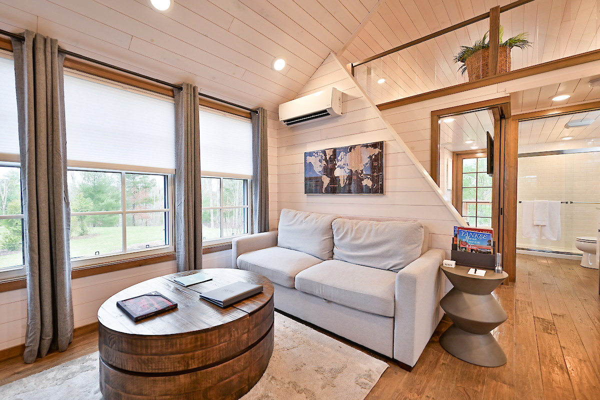 A cozy living room features a white sofa, round wooden coffee table, and large windows. The room is adjacent to a bathroom and a loft area.