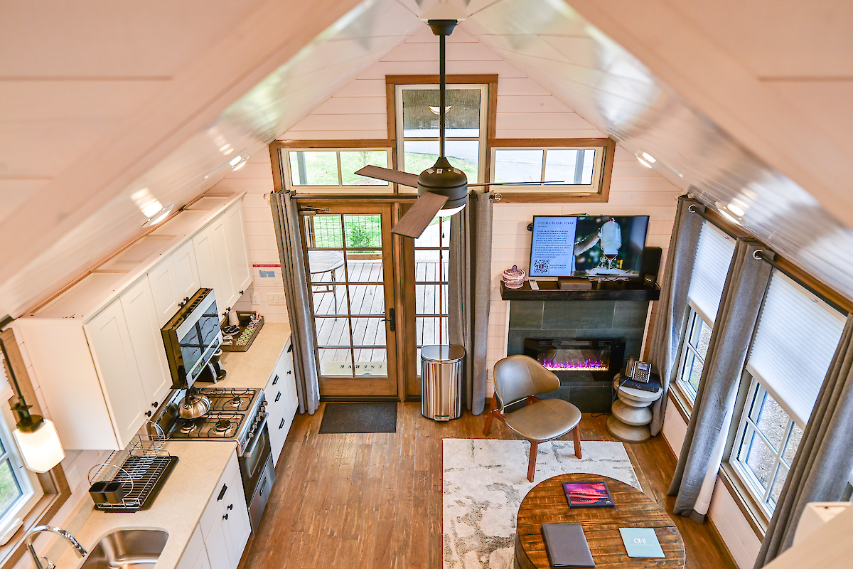 A cozy, small loft-style living space with a kitchen, couch, TV, and fireplace, viewed from above. There is also a ceiling fan and ample natural light.