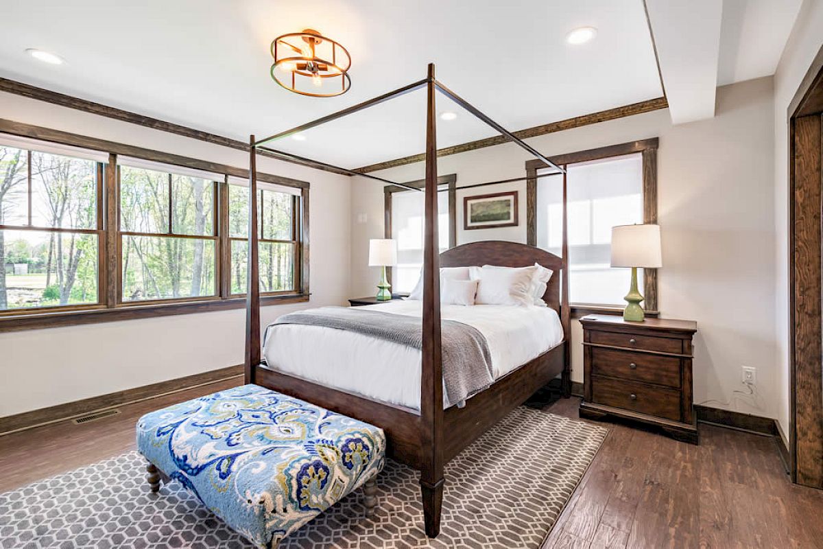 A cozy bedroom with a four-poster bed, two nightstands, a patterned ottoman, large windows, and modern lighting.