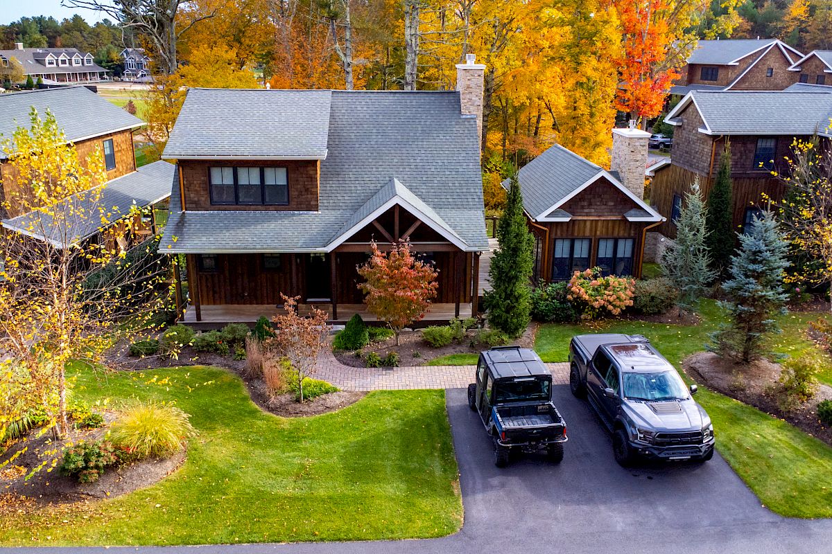 A cozy two-story house with a well-kept lawn, two vehicles parked in the driveway, surrounded by autumn foliage and neighboring houses.