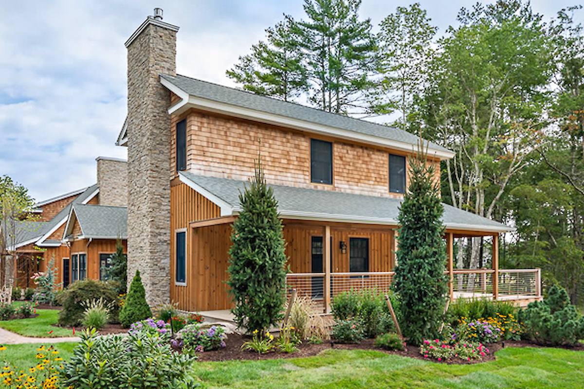 A two-story wooden house with a stone chimney, surrounded by trees, bushes, and a well-maintained garden with colorful flowers.