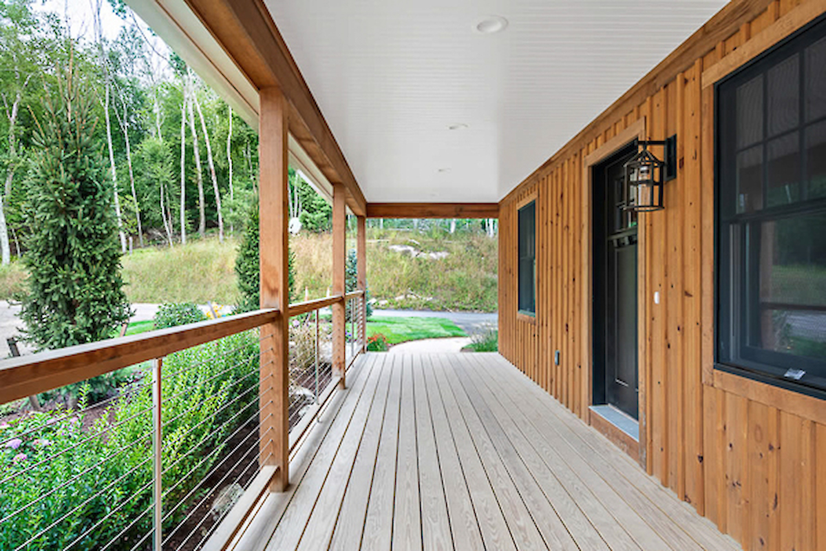 The image features a wooden porch with a ceiling and railing, attached to a house, overlooking a garden and surrounded by trees.