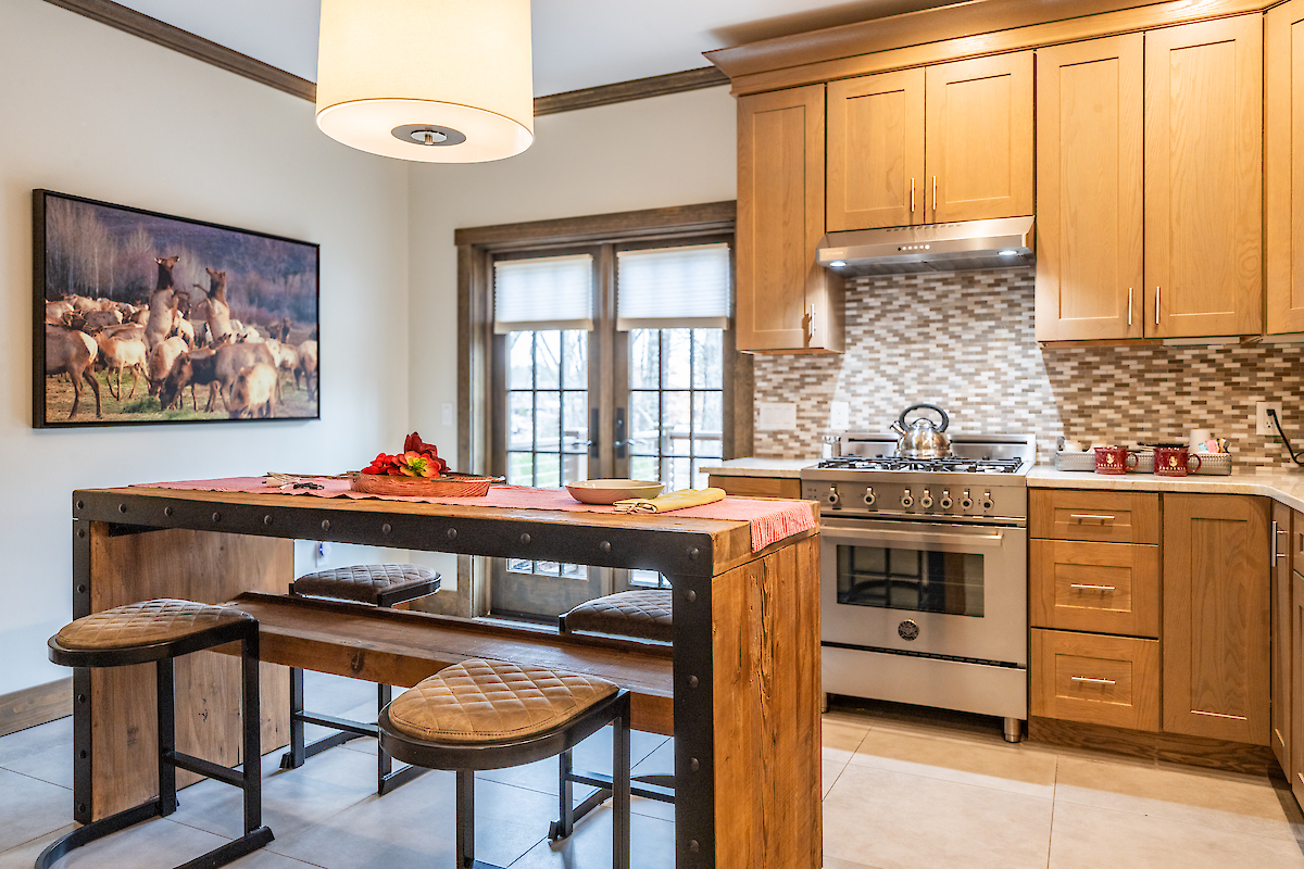 This image shows a modern kitchen with wooden cabinets, a dining table with stools, and a stove. A framed painting hangs on the wall.