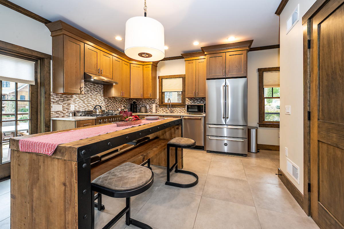 Modern kitchen with wooden cabinetry, stainless steel appliances, pendant light, island with bar seating, and a large window.