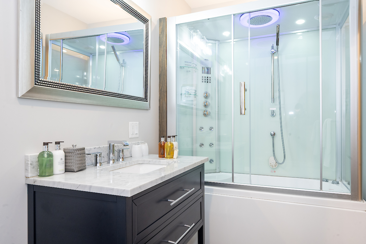 A modern bathroom features a sleek vanity with a marble countertop, a large mirror, and a glass-enclosed shower with advanced fixtures and lighting.