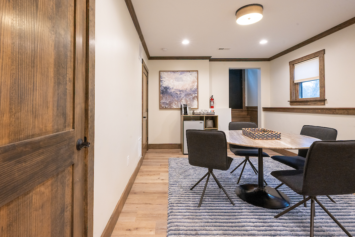 A modern room with a wooden door, a round table surrounded by four chairs, a textured rug, and a small counter with a fire extinguisher on it.