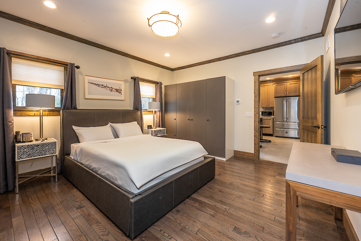A modern bedroom with a large bed, two nightstands, lamps, and a dresser. The room has hardwood floors and a connected doorway leading to a kitchen.