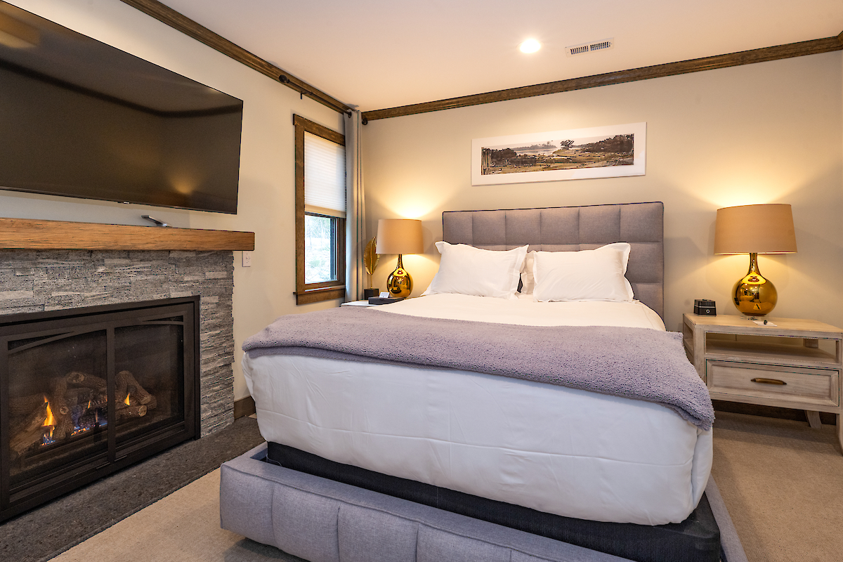 A cozy bedroom with a fireplace, TV, bed with white bedding, two bedside tables with lamps, and a scenic picture on the wall above the bed.