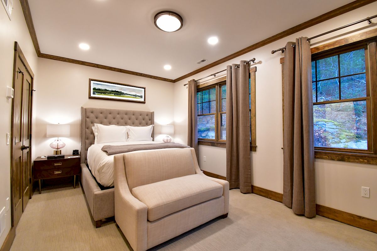 This image shows a cozy bedroom with a queen-sized bed, beige upholstered headboard, nightstands with lamps, a bench, and windows with beige curtains.