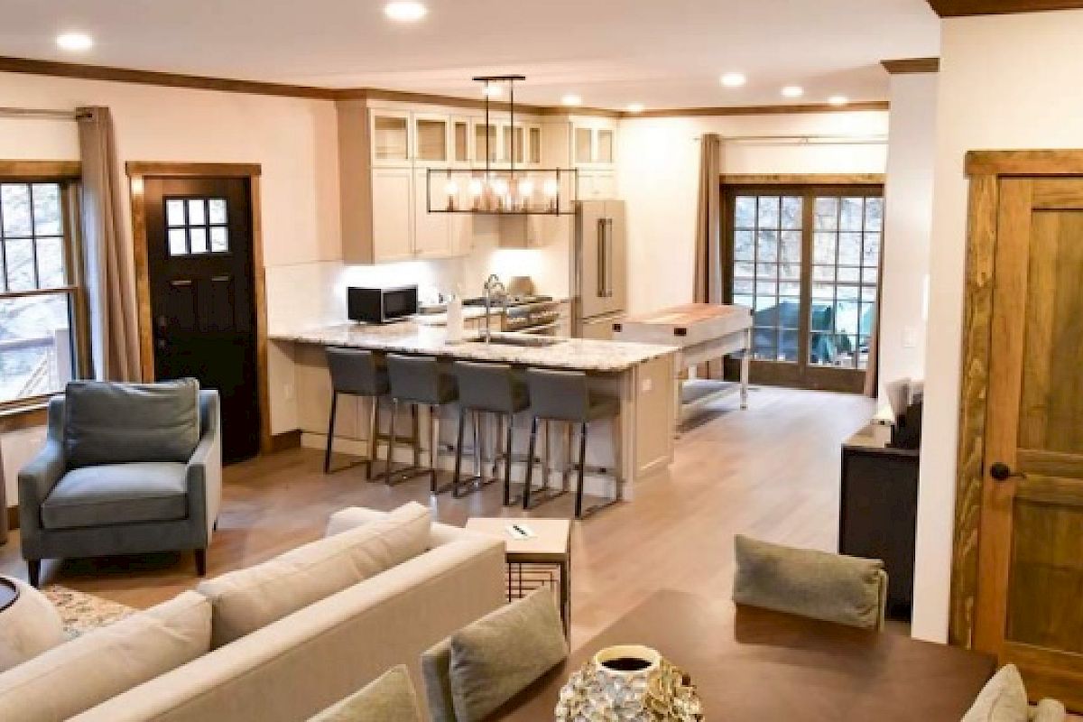 This image shows an open-concept living room and kitchen with a stone fireplace, modern furnishings, bar seating, and large windows and doors.