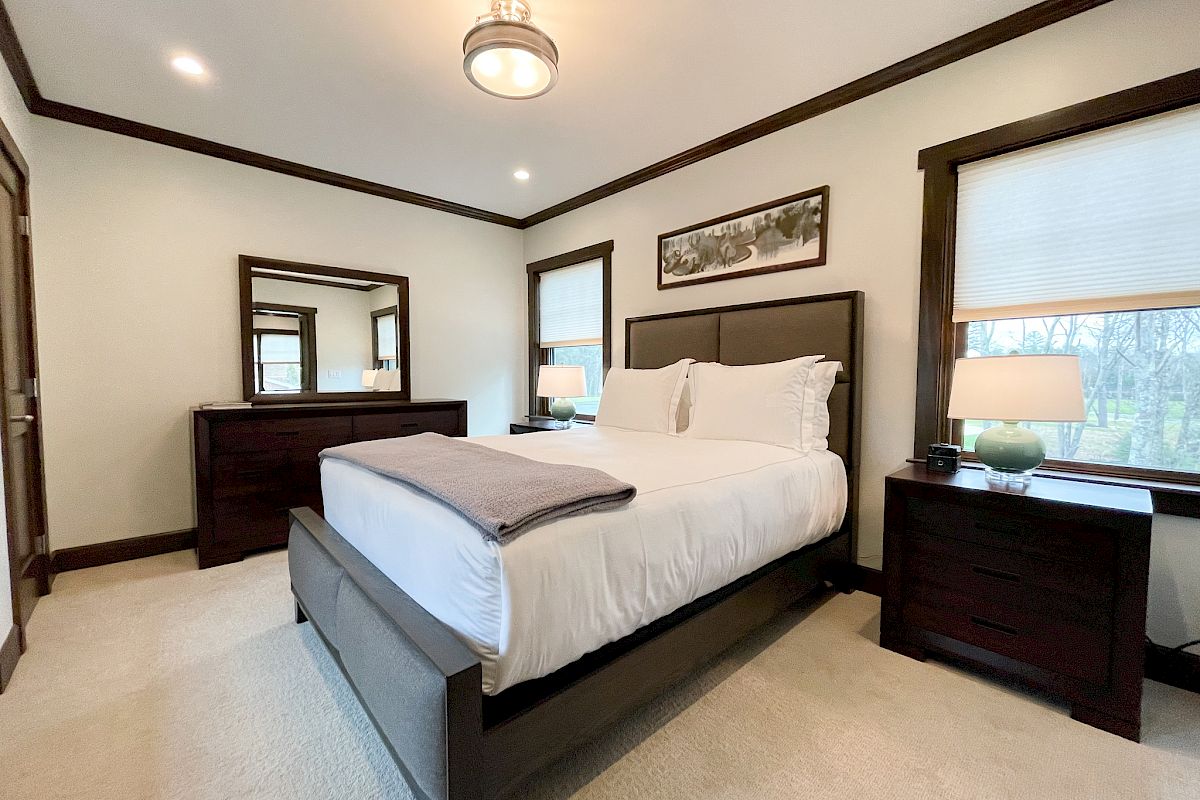 A modern bedroom with a bed, two side tables with lamps, a dresser with a mirror, clean white bedding, and wall art above the bed.