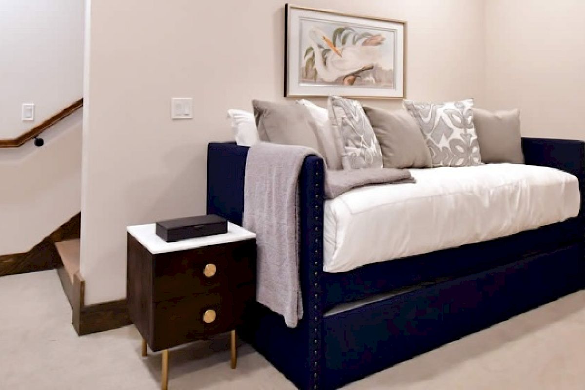 A cozy bedroom featuring a dark blue daybed with white and gray pillows, a nightstand with a drawer and book, and a framed artwork on the wall.