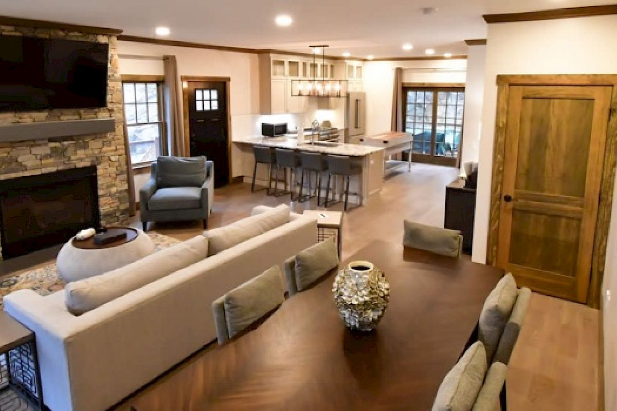 The image shows a modern, open-plan living area with a stone fireplace, kitchen with bar seating, dining table, and neutral-colored furniture.