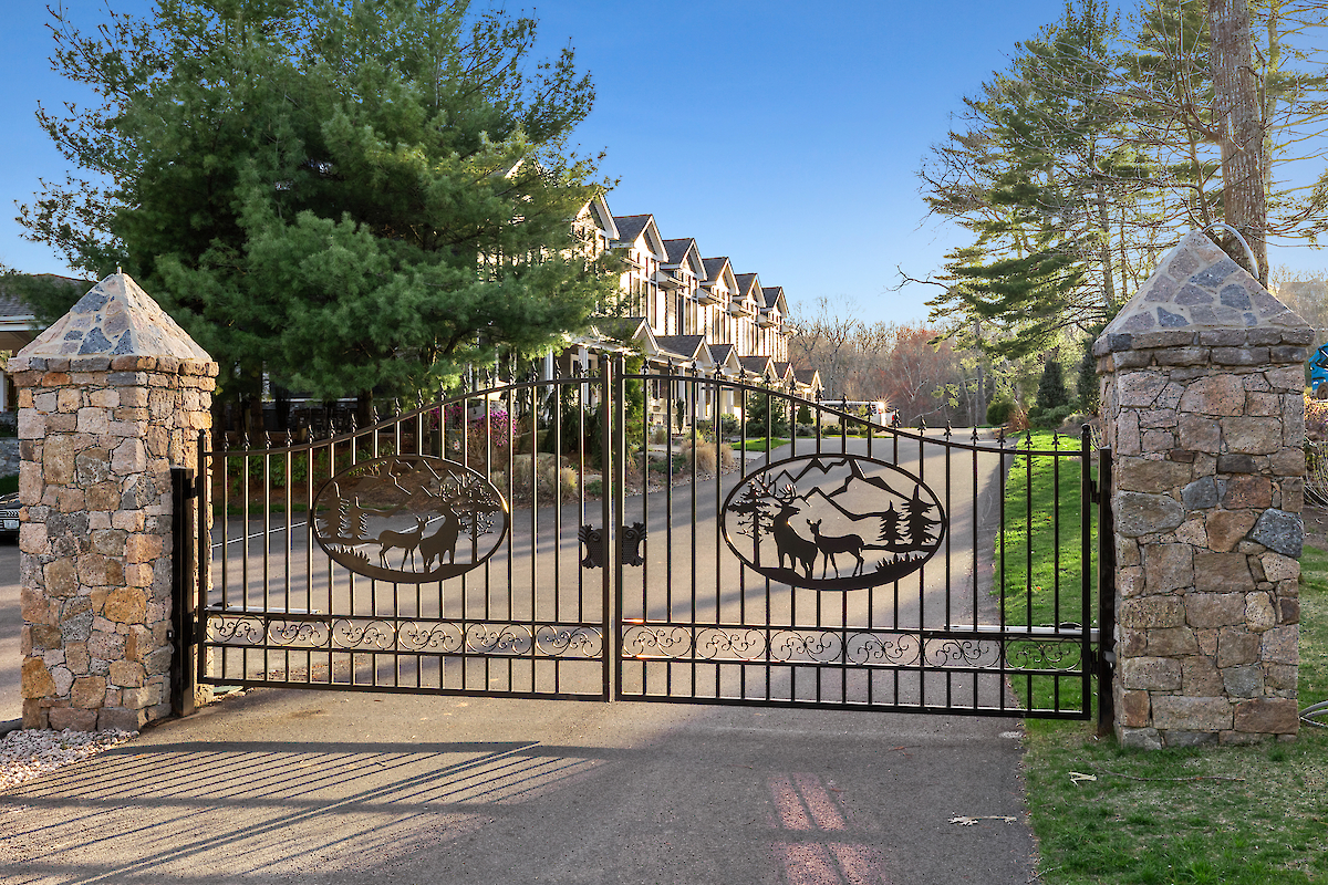 A wrought iron gate with decorative deer motifs, flanked by stone pillars, leading to a driveway lined with townhouses.