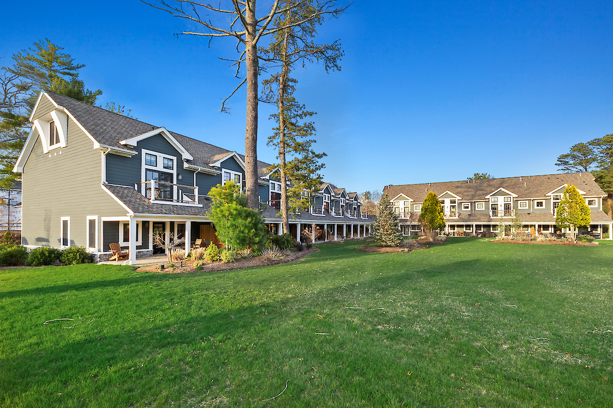 The image shows two multi-story buildings with balconies and a large grassy area in front, surrounded by trees under a clear blue sky.