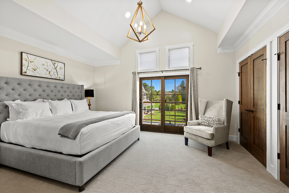A spacious bedroom with a large bed, upholstered headboard, armchair, and doors opening to a deck with a scenic view, featuring modern decor.