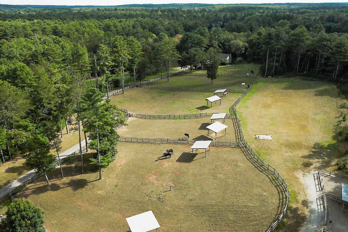 An aerial view of a spacious, fenced area with several shelters and surrounding forest. There are some people or animals within the fenced area, along paths.