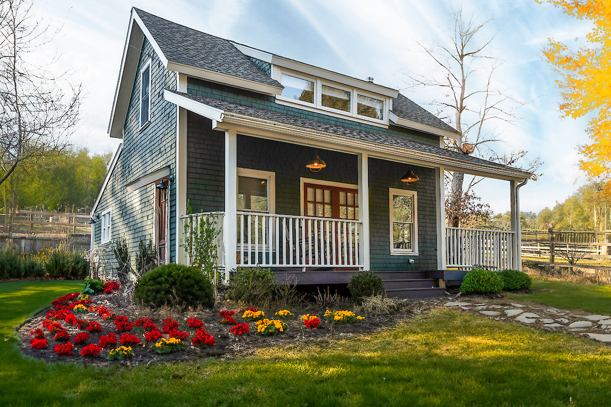 A charming blue house with a front porch, surrounded by a garden with red and yellow flowers, located in a rural setting.