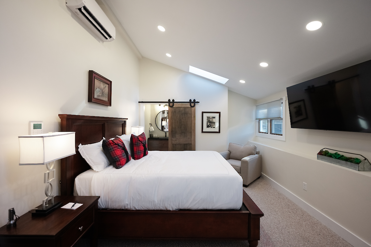 A modern bedroom with a large bed, decorative pillows, a ceiling with recessed lights, a barn door, wall-mounted TV, and a window.