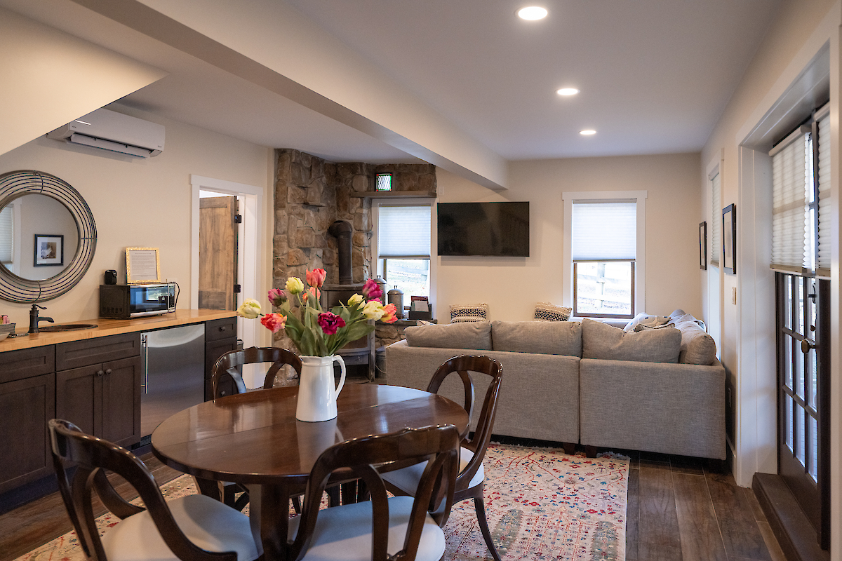 A cozy living and dining room features a round table, chairs, a large sofa, a TV, flowers in a vase, and a mix of modern and rustic decor.