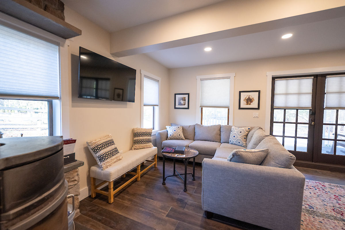 A cozy living room with a gray sectional sofa, two chairs, a wall-mounted TV, round table, framed artwork, and large windows with blinds ends the sentence.