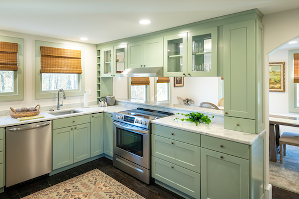 A modern kitchen with green cabinetry, stainless steel appliances, a rug, and a dining area visible through an archway in the background.