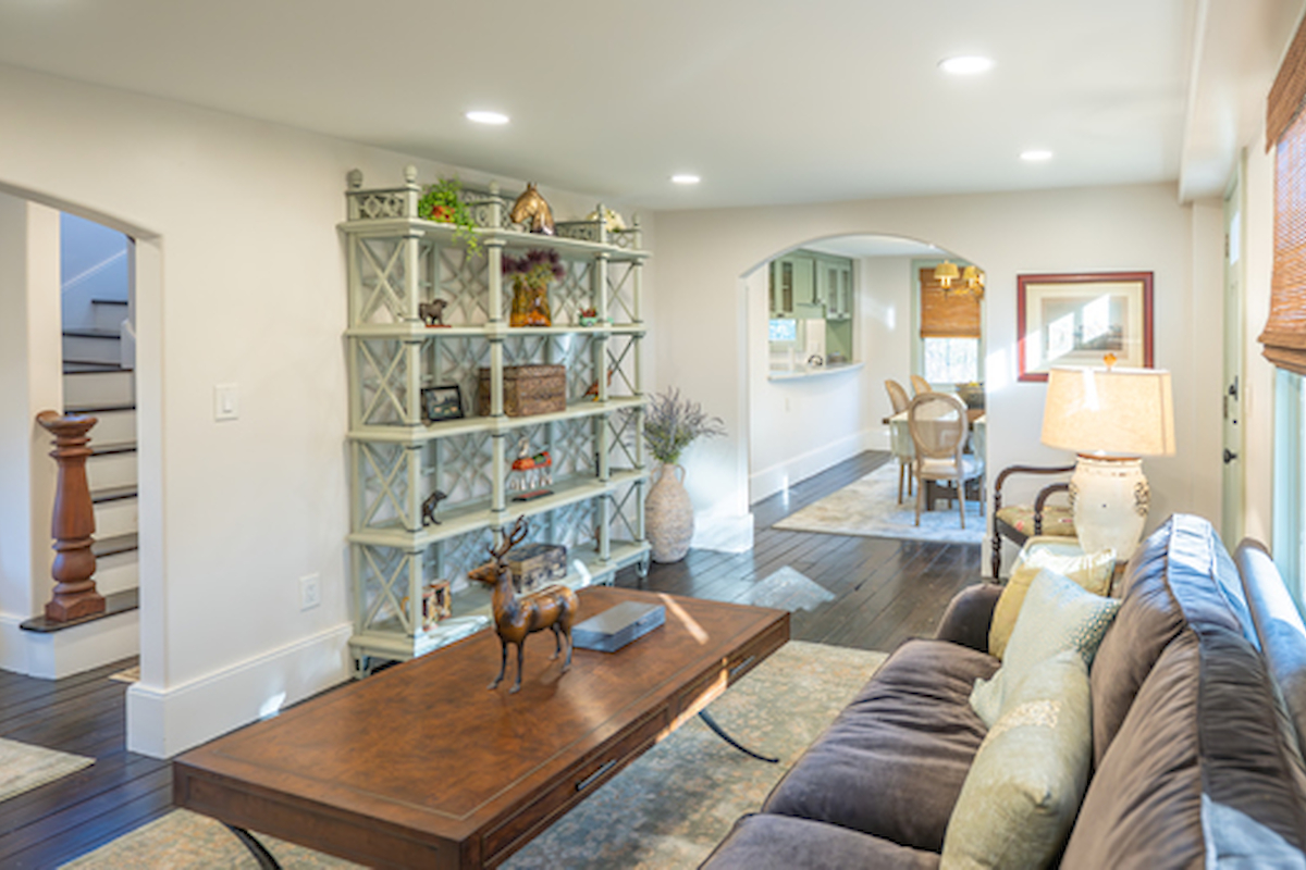 A cozy living room with a plush gray sofa, wooden coffee table, and open shelf, leading to a dining area under archways. Warm lighting throughout.