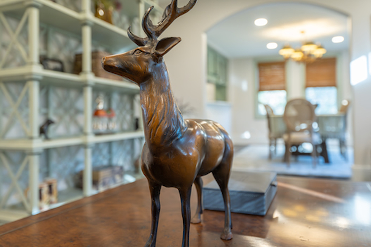 A bronze deer sculpture sits on a table in a living room, with a dining area and bookshelves in the background, featuring a cozy and bright ambiance.