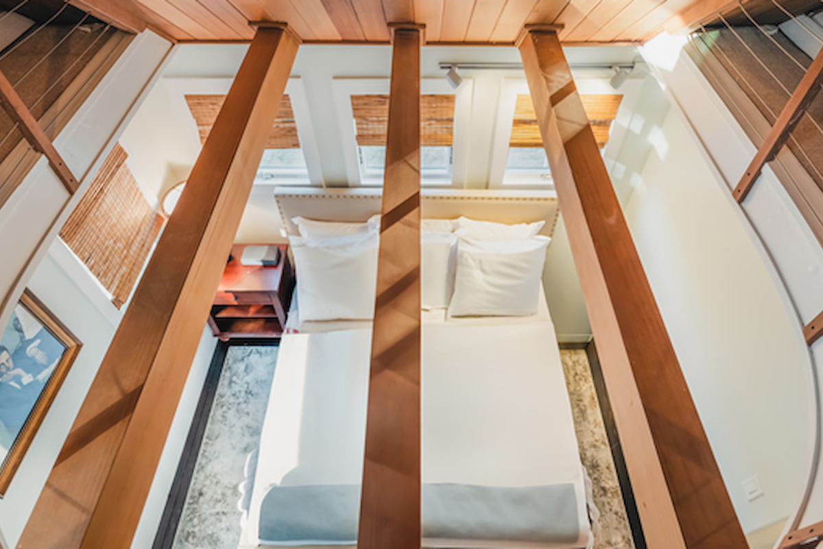 The image shows an overhead view of a narrow, well-lit bedroom with a bed, nightstand, and wooden beams across the ceiling.