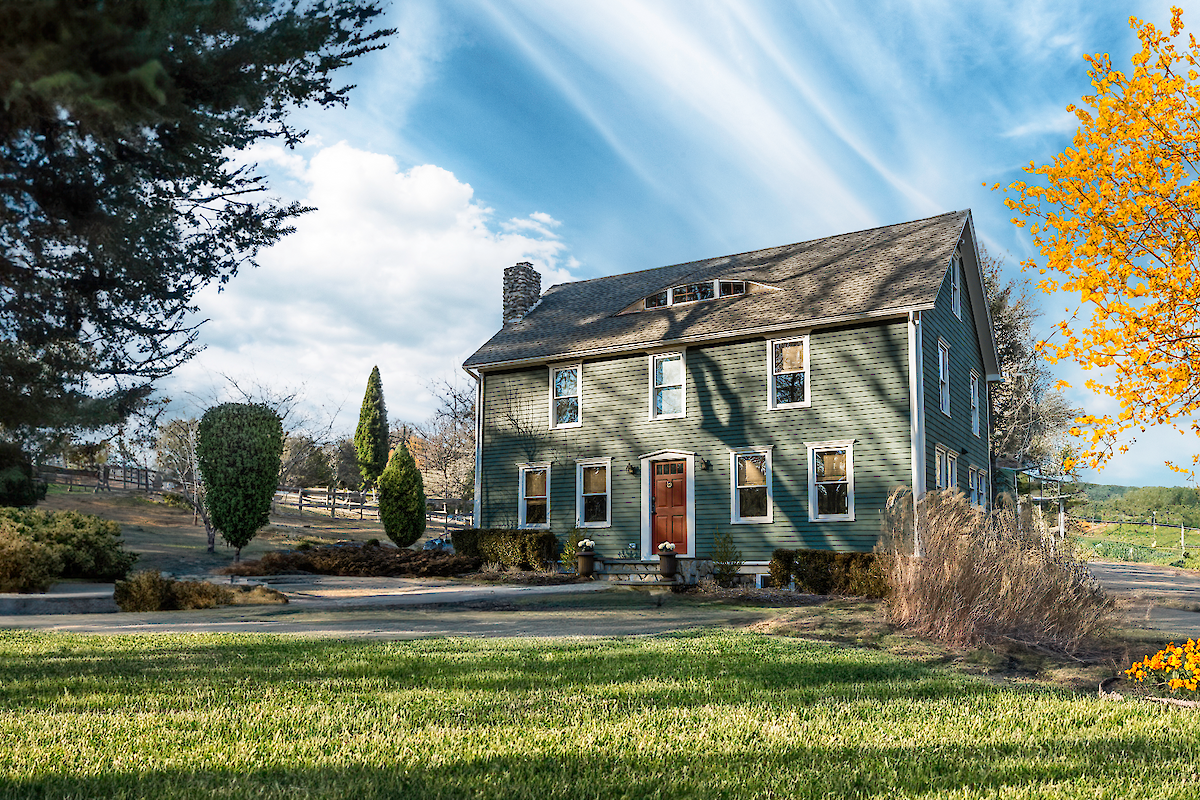 The image shows a green house with a sloped roof, surrounded by landscaped gardens, a tree with yellow leaves, and a clear sky with streaked clouds.