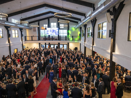 The image shows a formal event in a large hall with many people in suits and dresses, mingling and seated at tables.