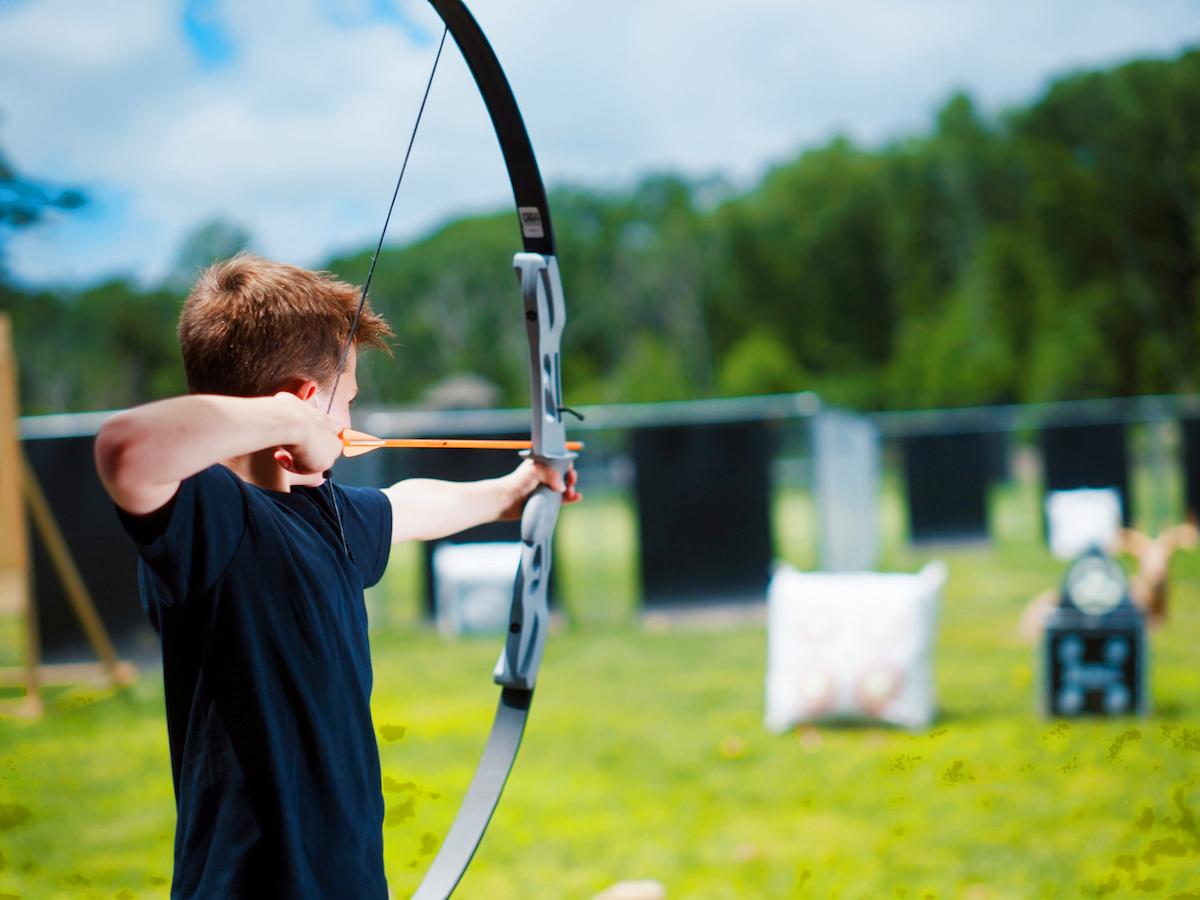 A person is aiming a bow and arrow at a target outdoors, with greenery and targets visible in the background.