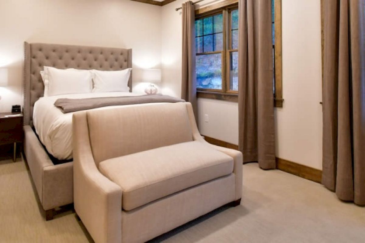 A cozy bedroom features a tufted bed, a bench at the foot, two nightstands with lamps, and windows with curtains.