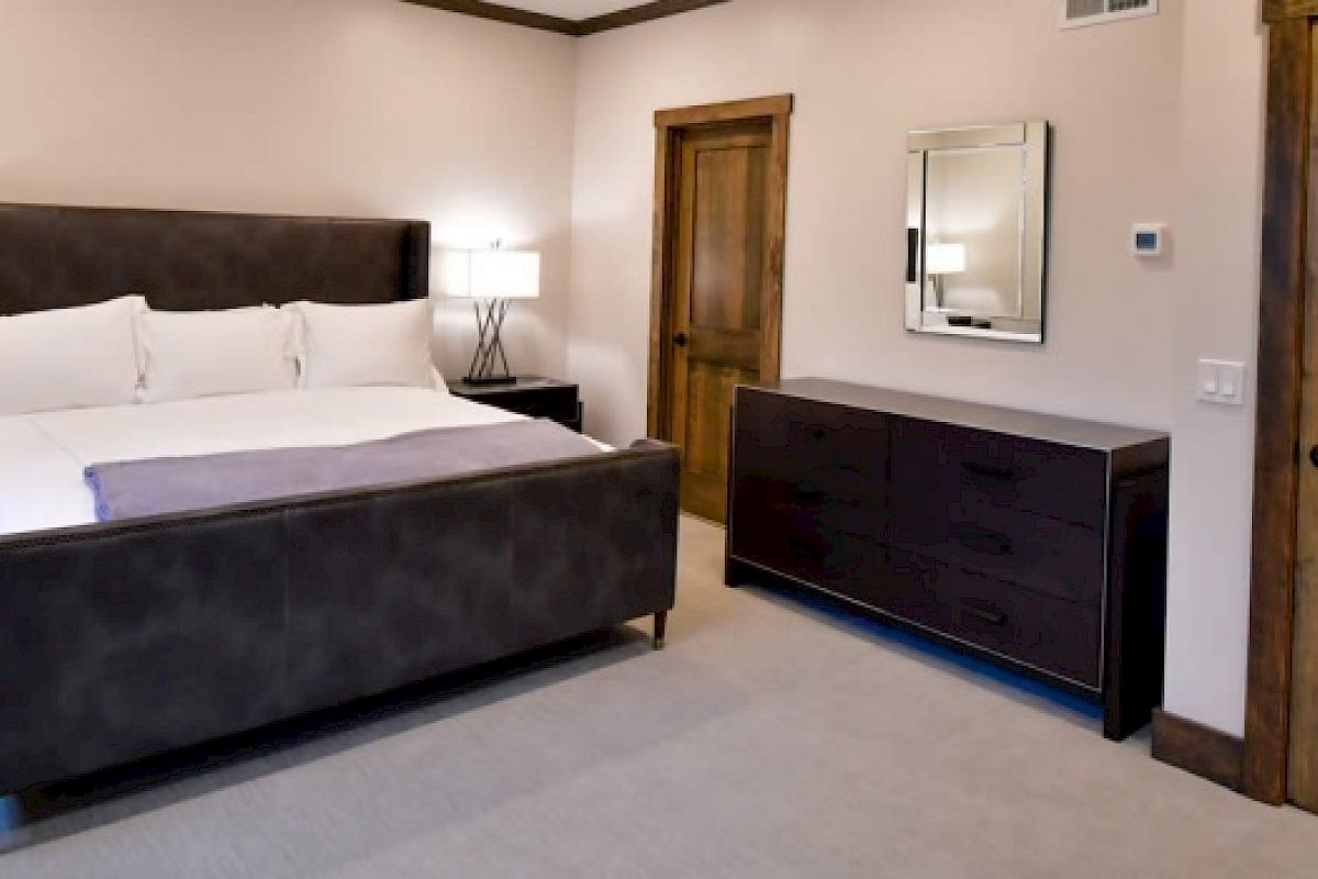 A bedroom with a large bed, bedside tables with lamps, a dresser, a mirror above the dresser, and two wooden doors, one open and one closed.