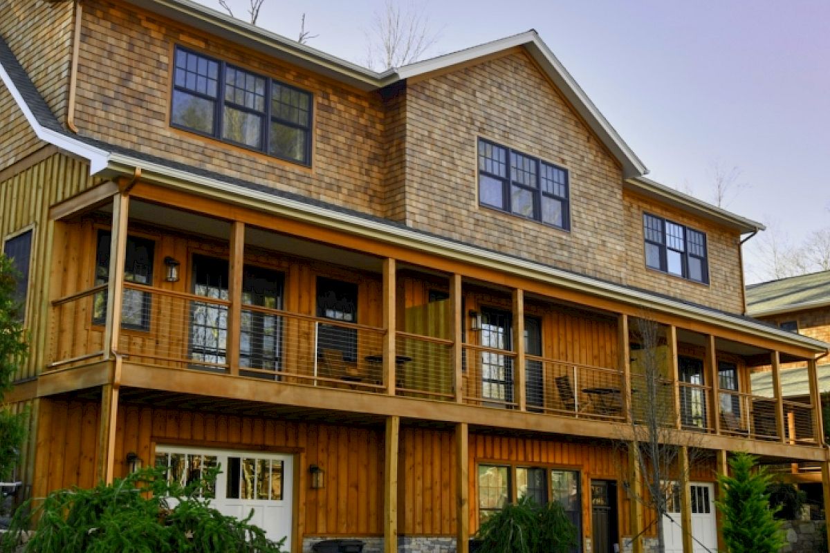 A large wooden house with two stories, large windows, and a spacious balcony is surrounded by greenery under a clear sky.