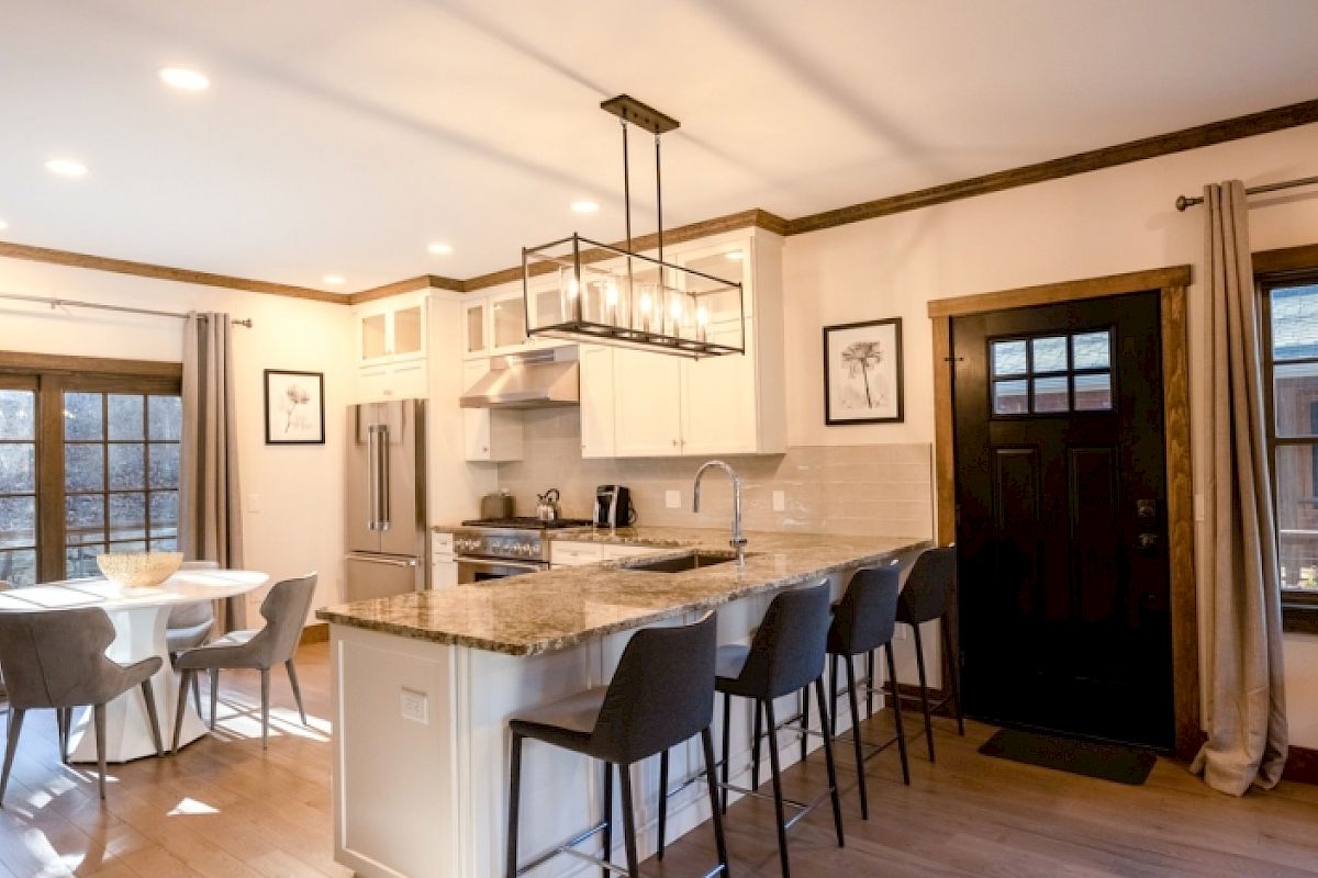 A modern kitchen and dining area with a white island, bar stools, dining table, large windows, and stainless steel appliances, ending the sentence.