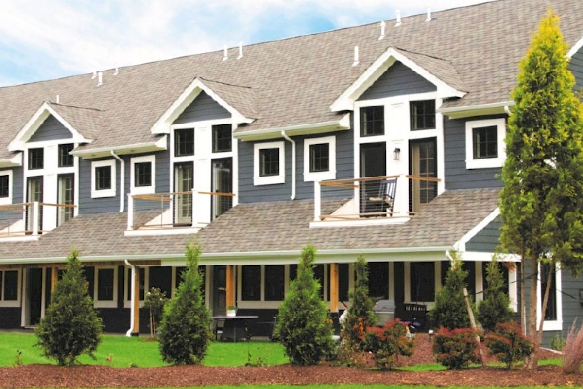 A row of modern townhouses with grey siding, white trim, porches, balconies, and a landscaped front yard featuring trees and shrubs.