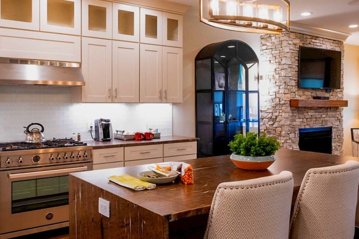 A modern kitchen with a large island, white cabinets, a stove, a brick archway leading to a cozy living room with a fireplace and TV.