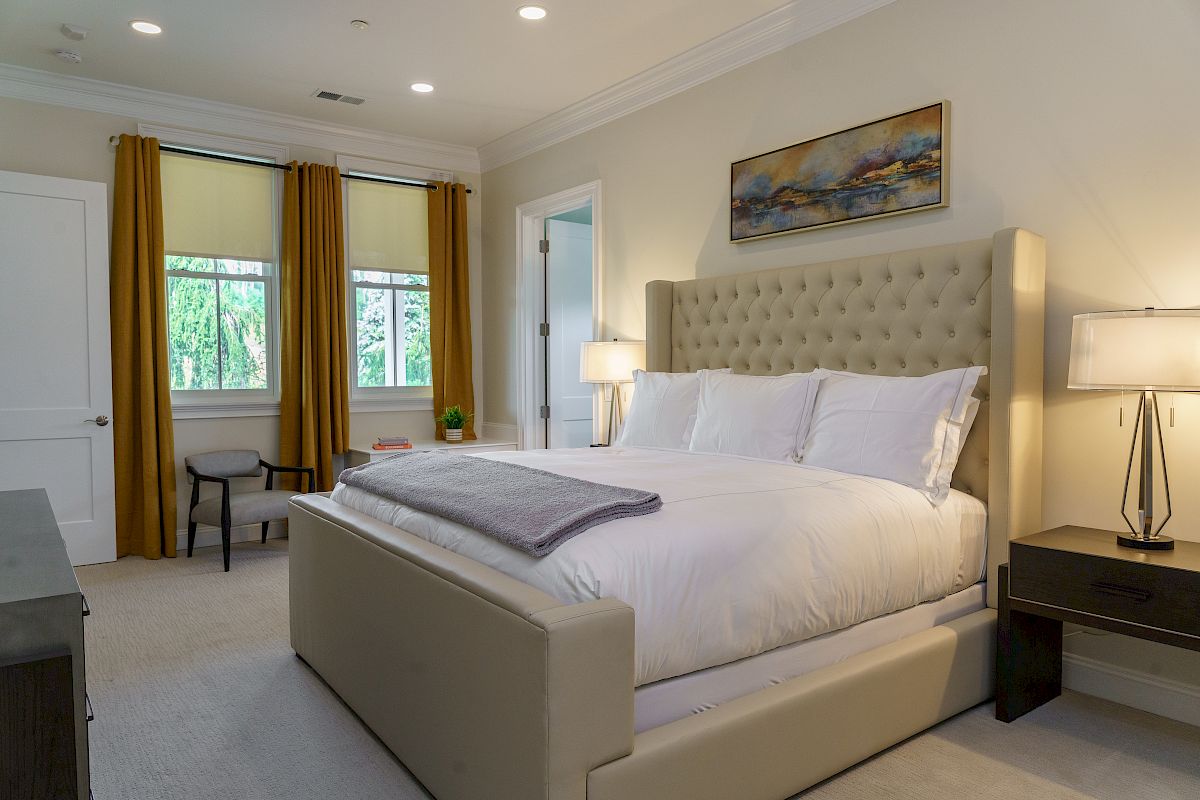 A modern bedroom with a large bed, white bedding, nightstands with lamps, a chair, golden curtains, and a landscape painting on the wall.