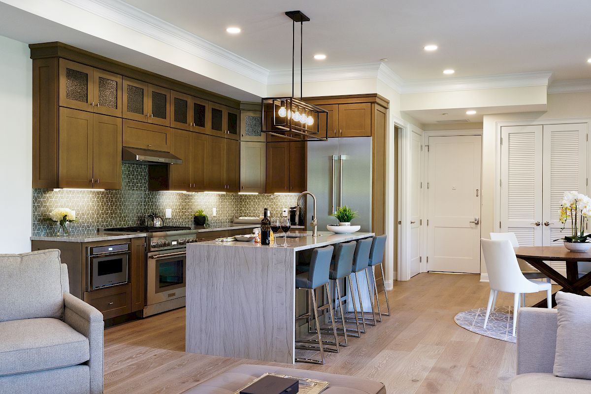 A modern kitchen with wooden cabinets, an island with barstools, stainless appliances, and an adjacent dining and living area is shown.