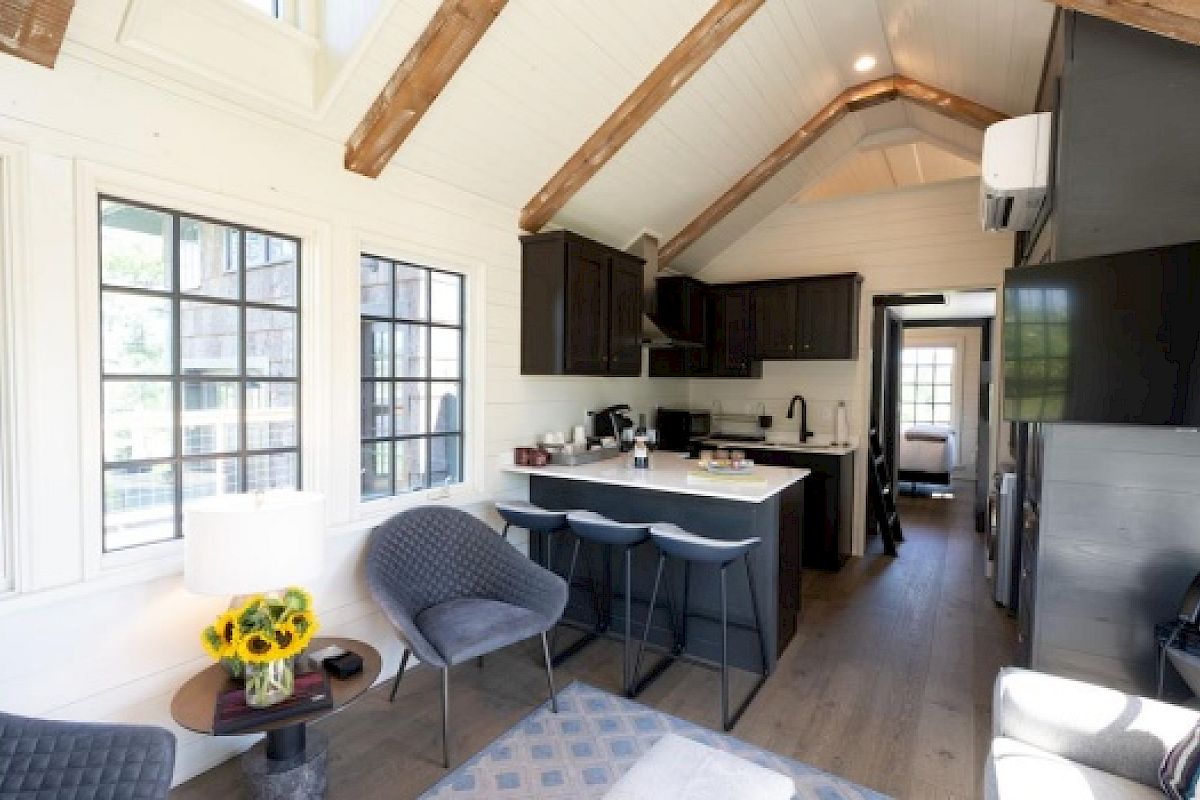 A cozy, modern tiny home interior with a kitchen, dining area, and sitting area. Exposed beams, dark cabinets, and a bouquet of sunflowers on a table end the sentence.
