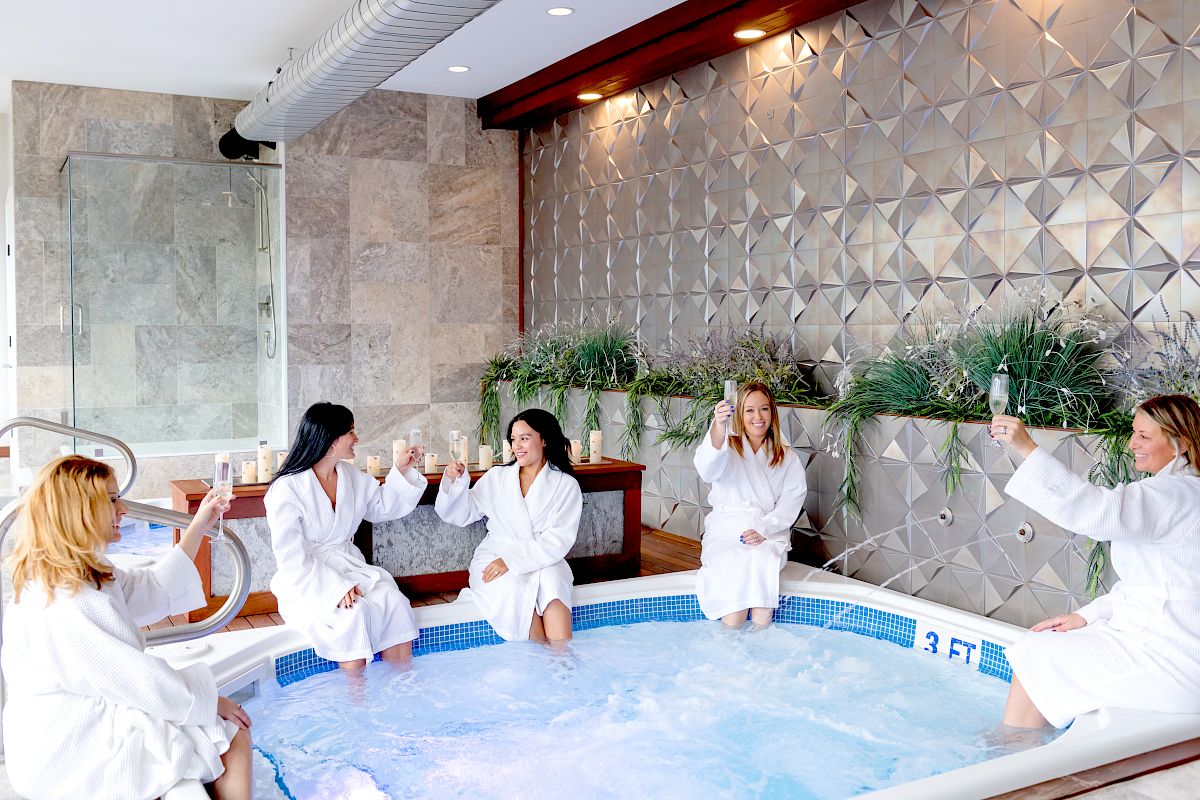 Five people in white robes are sitting around a hot tub, raising glasses in a toast. The space has modern decor and wall plants.