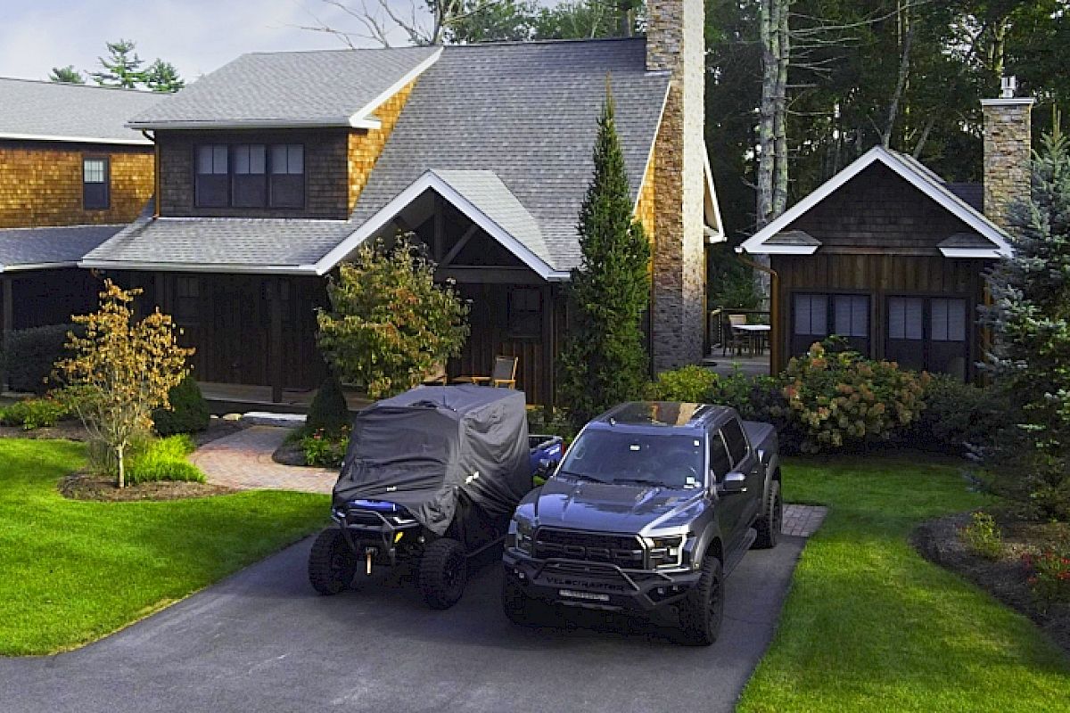A wooden house with a double garage, manicured lawn, a covered vehicle, and a black pickup truck in the driveway, surrounded by greenery.