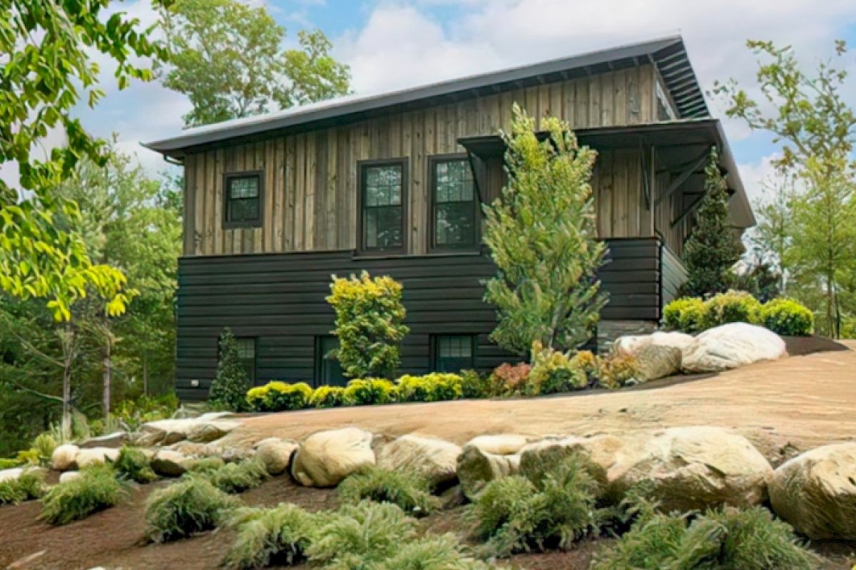 A modern wooden house with a slanted roof is surrounded by landscaping, including bushes, trees, and large rocks on a slightly elevated terrain.