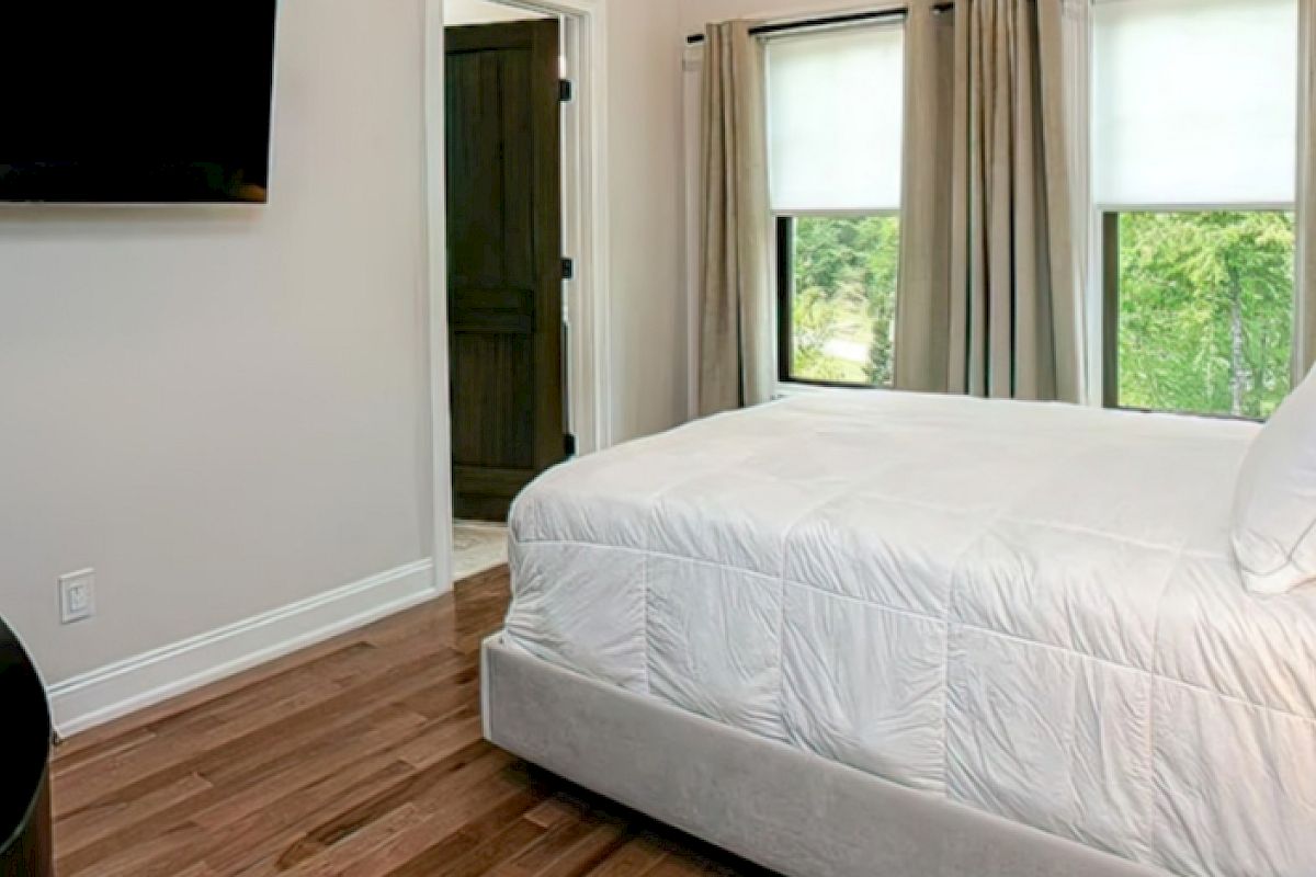A small bedroom with a neatly made bed, a wall-mounted TV, wooden flooring, and two windows with curtains and white blinds ending the sentence.