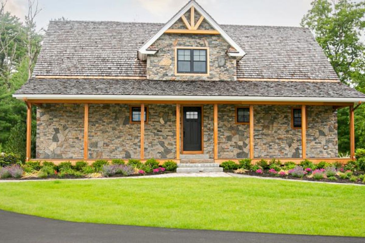 A stone house with a wooden roof surrounded by greenery, a manicured lawn, shrubs, and a paved driveway, under a cloudy sky.