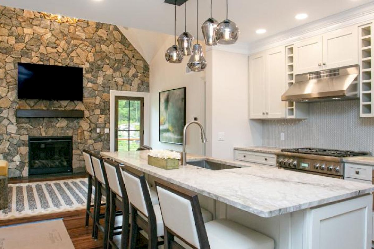 A modern kitchen and living room combo featuring a stone fireplace, TV, island with bar stools, and white cabinetry, ending the sentence.